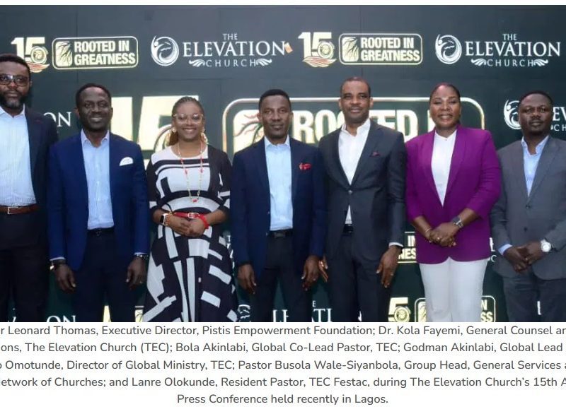 Group of seven well-dressed individuals posing on stage at The Elevation Church TEC 15th Anniversary press conference backdrop showing logos like Rooted in Greatness.